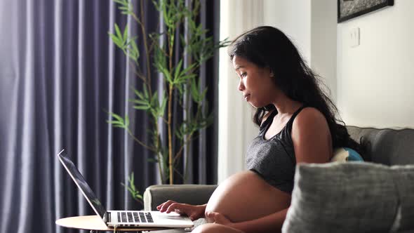 A Beautiful Pregnant Asian Woman Touching Belly While Working on Laptop at Home