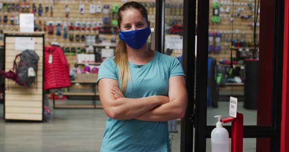 Caucasian female shopkeeper wearing face mask in the doorway of sports shop with arms crossed