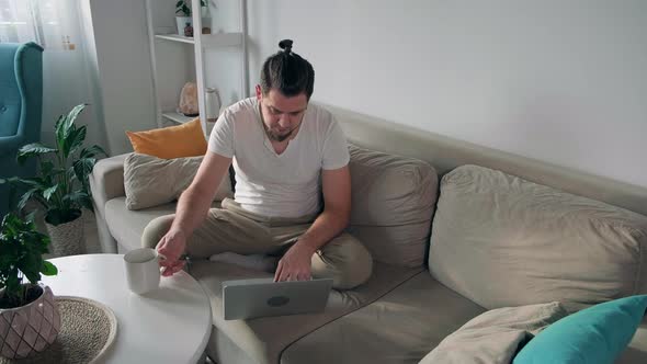 Young Man Using Laptop Typing and Working Sitting on the Couch at Home and Drink Coffee