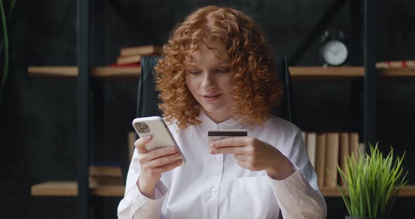 Pretty Redhaired Woman is Making Online Payment Holding Bank Credit Card Using Smartphone at Office