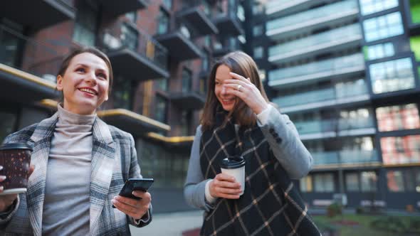 Two Happy Women Walking with Takeaway Coffee and Talking with Interest Among Themselves in the