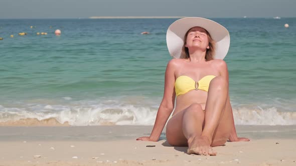 Portrait Young Smiling Woman Sitting on Beach in Yellow Swimsuit and White Hat