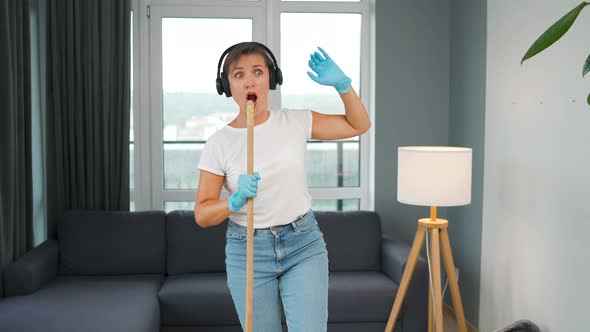 Woman in Headphones Cleans the House and Have Fun Singing with a Broom Like a Star at a Concert
