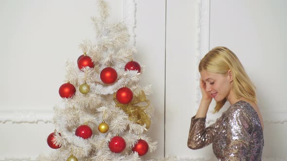 Happy Young Blonde Girl Decorating Christmas Tree. Young Woman Holding Bauble in Front of Christmas