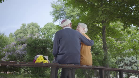 Back of Old Man Sit in the Bench in the Park with Bouquet of Yellow Flowers and Hug Woman