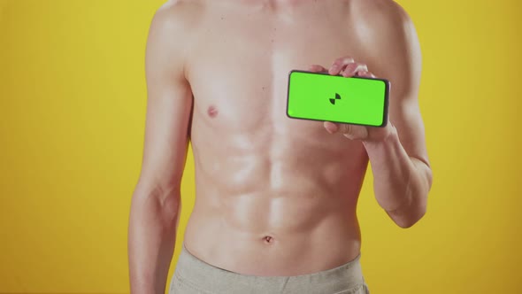 Abs of an athletic man on yellow background in studio raises phone with green screen in hand