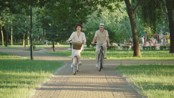 Wide Shot of Joyful Mid-adult Man and Woman Riding Bikes in Summer Park at Sunset. Portrait of Happy