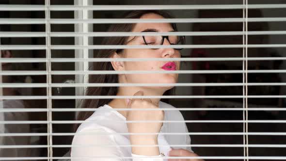Young Woman Sits at the Window View Through the Blinds