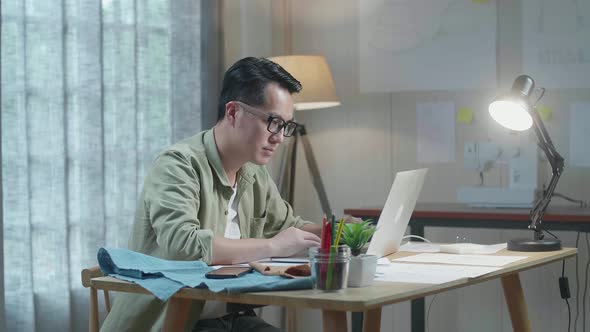 Side View Of Asian Man Designer With Layout Bond, Fabric, And Smartphone Working With Laptop