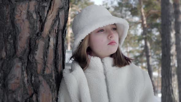 Portrait Thinking Young Woman in White Hat and Fur Coat Leaning to Tree Trunk in Winter Forest