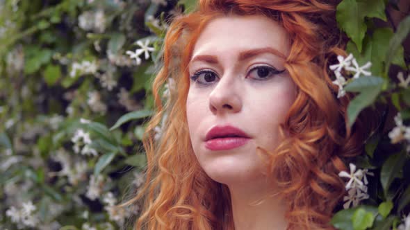 pensive red-haired woman among the jasmine stares at camera with puzzled look