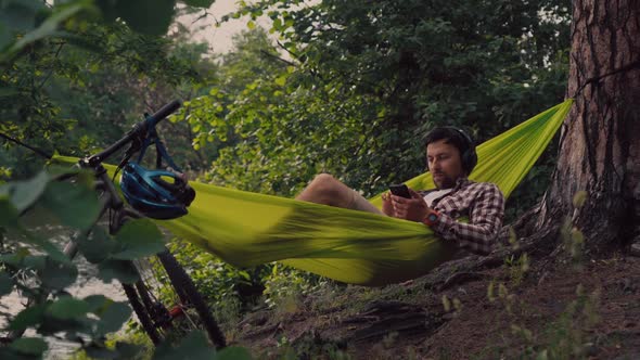 Man Travels on Bicycle Relaxing in Green Hammock Surfing Internet on Smartphone Listening Music on