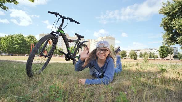 12 Year Old Girl Lying on the Grass Next to Her Bike and Waving Her Hand