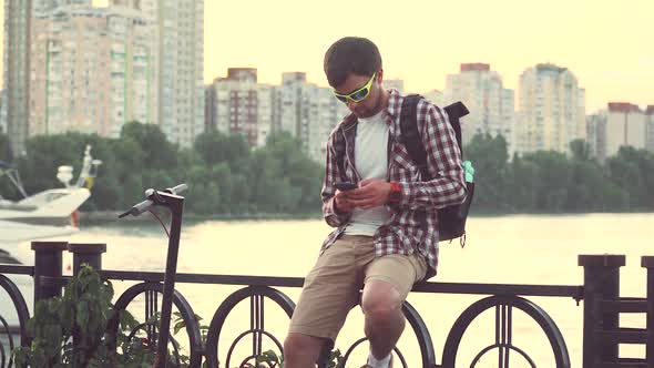 Young Caucasian Man Sitting on Fence on River Embankment Uses Smartphone Chatting While Waiting for