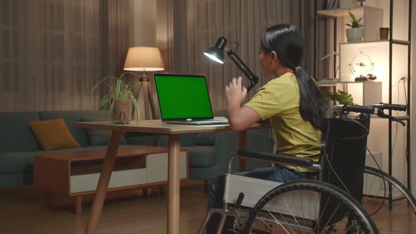 Asian Kid Girl Sitting In A Wheelchair And Raise Hand While Learning Online On Laptop Green Screen