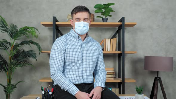 Serious businessman wears protective medical mask on face sits at desk in office