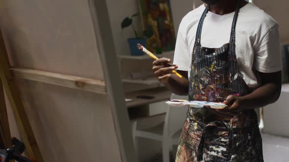 Thoughtful african american male artist holding paint brushes and palette at art studio