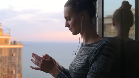 Smiling Girl Standing By the Window During a Sunny Day and Listening to Music with Headphones Using