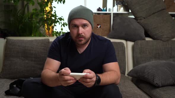 Young Attractive Bearded Guy in Gray Hat Sitting on the Couch and Playing Console. Very Happy Win.