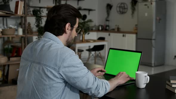 Young man using green screen chroma key laptop typing working in light office with modern computer