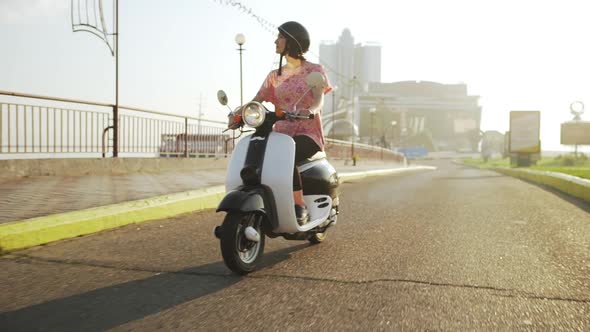 Young Beautiful Girl Riding Around City on Scooter at Sunrise