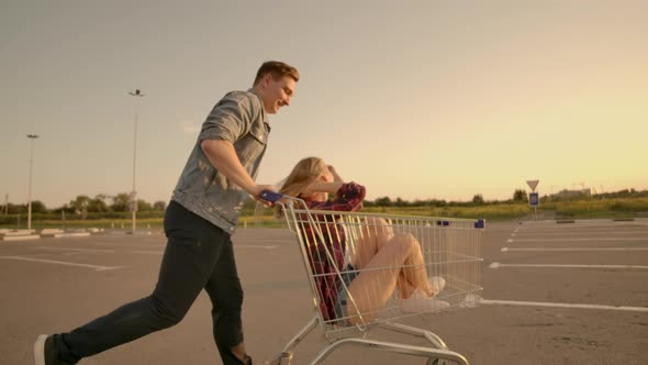 Cheerful People a Couple a Man and a Woman at Sunset Ride on Supermarket Trolleys in Slow Motion