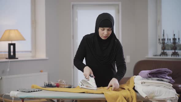 Portrait of Concentrated Muslim Woman in Hijab Ironing Yellow Pullover. Housewife Doing Housework