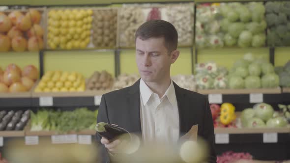 Portrait of Young Confident Caucasian Man Choosing Eggplant in Grocery and Putting Vegetable Into