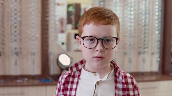 A Redhaired Caucasian Boy in a Plaid Red and White Shirt Stands with Transparent Glasses for Vision