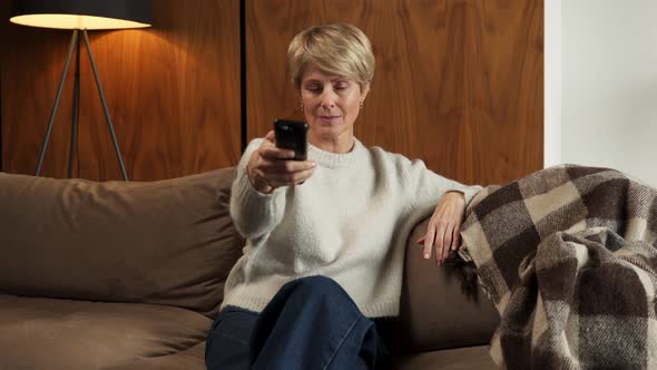 Smiling Middleaged Woman Sitting on the Sofa and Turning on the TV with a Remote Control