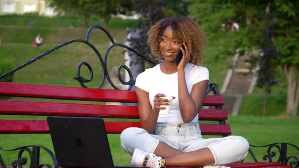 Happy African Woman Talking on the Phone While Sitting on a Bench in the Park and Drinks Coffee