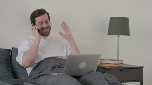 Young Man with Laptop Talking on Smartphone in Bed