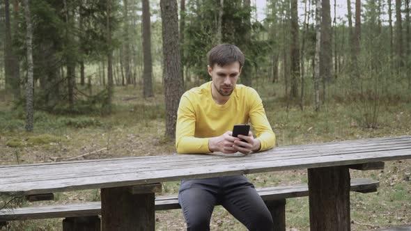 Man with Stubble Sits on Wooden Bench at Table in Forest Looks at Smartphone