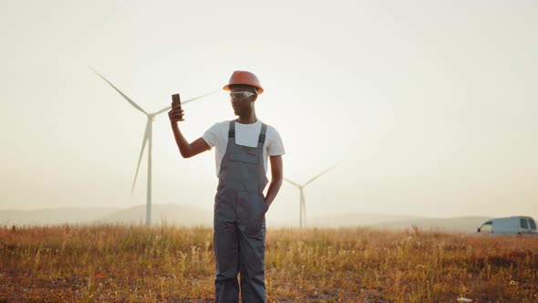 Engineer in Uniform Using Modern Smartphone for Video Call Among Windmill Farm