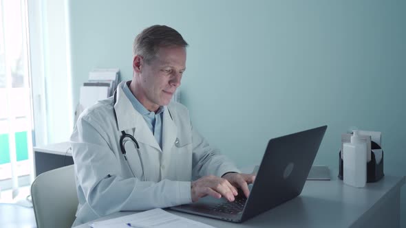 Smiling Old Doctor Using Laptop Computer Sitting at Desk in Medical Office