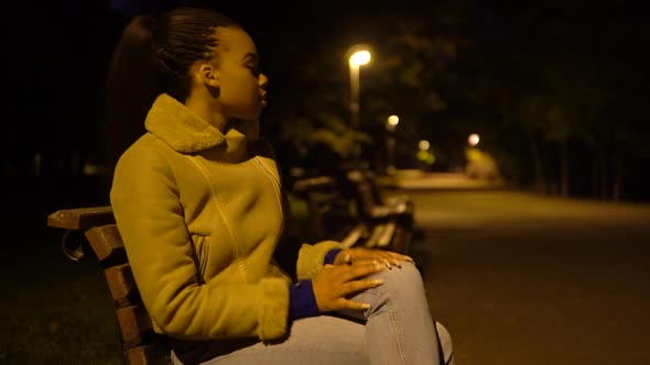 A Young Black Woman is Frustrated As She Sits on a Bench in a City Park at Night  Side View