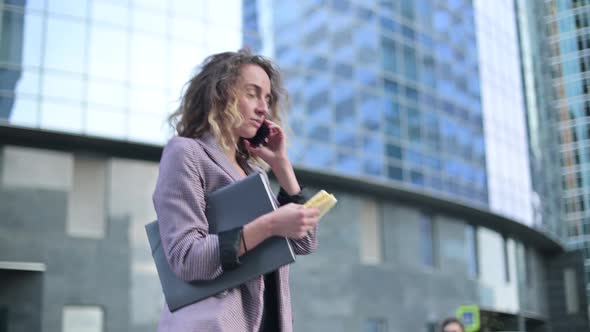 Side view of young business woman eating on the go and talking on mobile phone