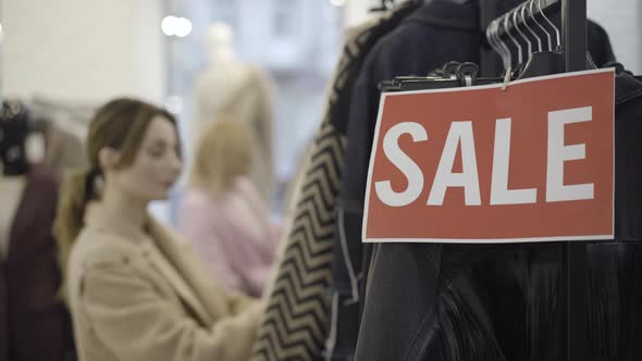 Close-up of Sale Banner on Pricey Clothes Hanging on Rack with Blurred Beautiful Caucasian Women