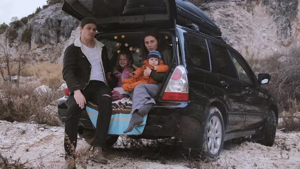 Traveler Family  sitting in the open trunk of a black car with kids near bonfire