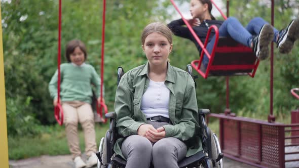 Medium Shot Sad Paraplegic Girl in Wheelchair Looking at Camera Sighing As Friends Swinging on