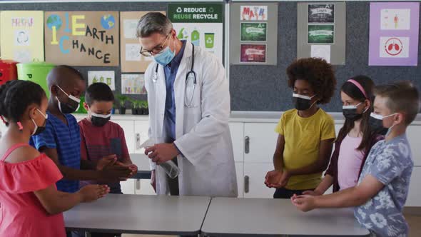 Diverse schoolteacher and schoolchildren standing disinfecting hands, all wearing face masks