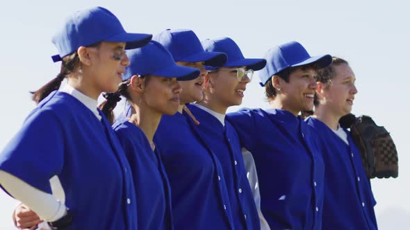 Happy diverse team of female baseball players standing in line with arms around each other singing