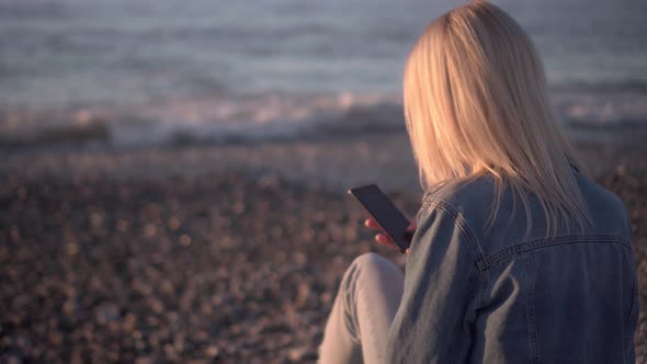 blonde in denim suit is sitting on seashore at sunset with smartphone in hands