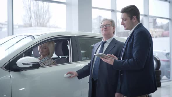 Beautiful Mature Caucasian Blond Woman Sitting in Car As Her Senior Husband Talking with Car Dealer