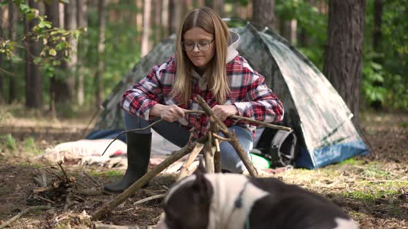 Panning Shot of Concentrated Young Woman Setting Camp Fire Breaking Branches with Curios Dog Resting