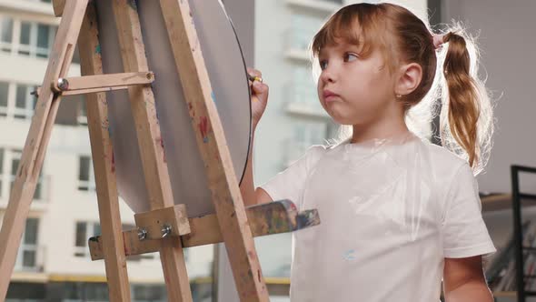 Beautiful Light Hair Little Girl in Transparent Apron Draws with Paintbrush on Canvas on an Easel