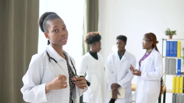 A Young African American Female Doctor Stands in Front and Looks at the Camera Against the