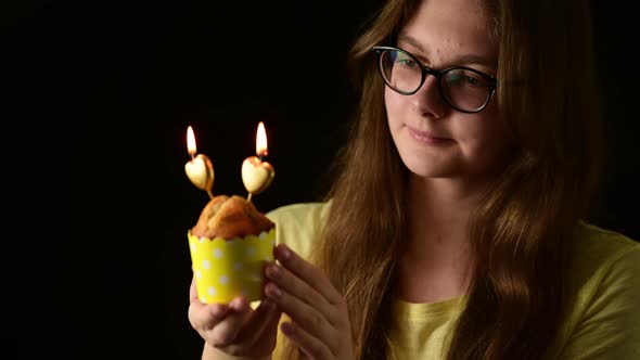 Happy attractive young woman holding one muffin with two heart shape candle, blows out the candles