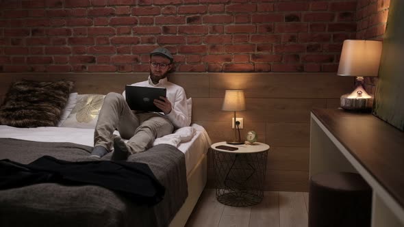 Handsome young businessman in a formal white shirt sits on a bed with a tablet in a modern room.