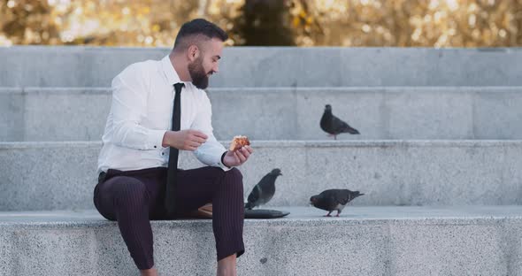 Happy Mature Businessman Feeding Pigeons with Pizza in Autumn Urban Park